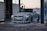 A person is washing a silver BMW car with high-pressure water at a car wash station. The car is covered in soap suds, and the sun appears to be setting or rising in the background.