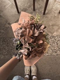 A person holding a bouquet of flowers wrapped in a light brown paper. The bouquet consists of various flowers including pink and purple blossoms. The focus is on the detailed textures and colors of the flowers. The background features a tiled floor with chairs, giving a casual indoor setting.