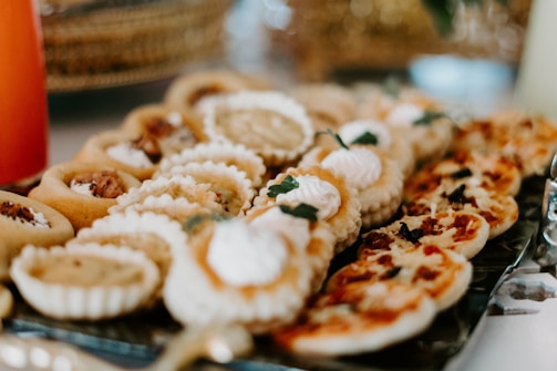 A close-up of a tray filled with an assortment of small pastries and savory tarts, some topped with whipped cream or garnished with herbs. Various textures and shapes are visible, including flaky crusts and creamy fillings, suggesting a variety of flavors.