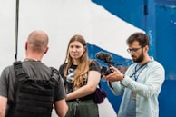 Three people are visible; one person is holding a video camera with an attached microphone, focusing on the other two individuals. The person being filmed has their back to the camera and is wearing a black harness or vest, while the other person stands facing the camera with a neutral expression. The background shows a white and blue wall, likely outdoors.