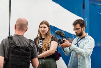 Three people are visible; one person is holding a video camera with an attached microphone, focusing on the other two individuals. The person being filmed has their back to the camera and is wearing a black harness or vest, while the other person stands facing the camera with a neutral expression. The background shows a white and blue wall, likely outdoors.
