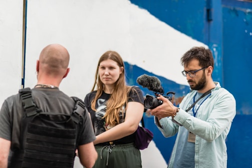 Three people are visible; one person is holding a video camera with an attached microphone, focusing on the other two individuals. The person being filmed has their back to the camera and is wearing a black harness or vest, while the other person stands facing the camera with a neutral expression. The background shows a white and blue wall, likely outdoors.