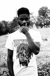 A person stands outdoors dressed in a graphic t-shirt with intriguing artwork on the front. The scene is set in a grassy field with trees in the distance, giving off a natural and serene atmosphere. The image is in black and white, enhancing the somber yet artistic feel.