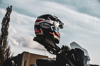 A motorcycle helmet with a pattern of red, white, and black colors is perched on top of a motorcycle against a backdrop of a clear sky. The angle suggests an outdoor setting with a cloudy sky and a tall building partially visible.