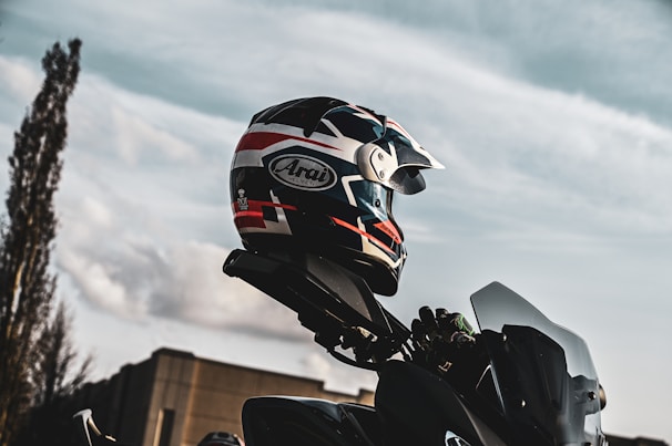 A motorcycle helmet with a pattern of red, white, and black colors is perched on top of a motorcycle against a backdrop of a clear sky. The angle suggests an outdoor setting with a cloudy sky and a tall building partially visible.