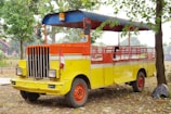 A brightly colored, open-air vehicle with a vivid combination of yellow, orange, and red hues. The vehicle has a retro design with a grille and two large headlights at the front. It is parked on a patch of dry ground, surrounded by trees and scattered leaves. An Indonesian flag is attached to the front, adding a cultural element to the scene.