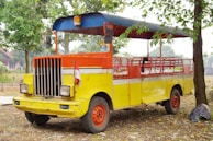 A brightly colored, open-air vehicle with a vivid combination of yellow, orange, and red hues. The vehicle has a retro design with a grille and two large headlights at the front. It is parked on a patch of dry ground, surrounded by trees and scattered leaves. An Indonesian flag is attached to the front, adding a cultural element to the scene.