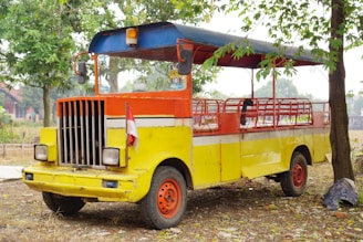 A brightly colored, open-air vehicle with a vivid combination of yellow, orange, and red hues. The vehicle has a retro design with a grille and two large headlights at the front. It is parked on a patch of dry ground, surrounded by trees and scattered leaves. An Indonesian flag is attached to the front, adding a cultural element to the scene.