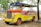 A brightly colored, open-air vehicle with a vivid combination of yellow, orange, and red hues. The vehicle has a retro design with a grille and two large headlights at the front. It is parked on a patch of dry ground, surrounded by trees and scattered leaves. An Indonesian flag is attached to the front, adding a cultural element to the scene.