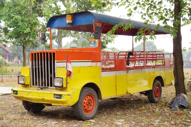 A brightly colored, open-air vehicle with a vivid combination of yellow, orange, and red hues. The vehicle has a retro design with a grille and two large headlights at the front. It is parked on a patch of dry ground, surrounded by trees and scattered leaves. An Indonesian flag is attached to the front, adding a cultural element to the scene.