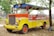 A brightly colored, open-air vehicle with a vivid combination of yellow, orange, and red hues. The vehicle has a retro design with a grille and two large headlights at the front. It is parked on a patch of dry ground, surrounded by trees and scattered leaves. An Indonesian flag is attached to the front, adding a cultural element to the scene.