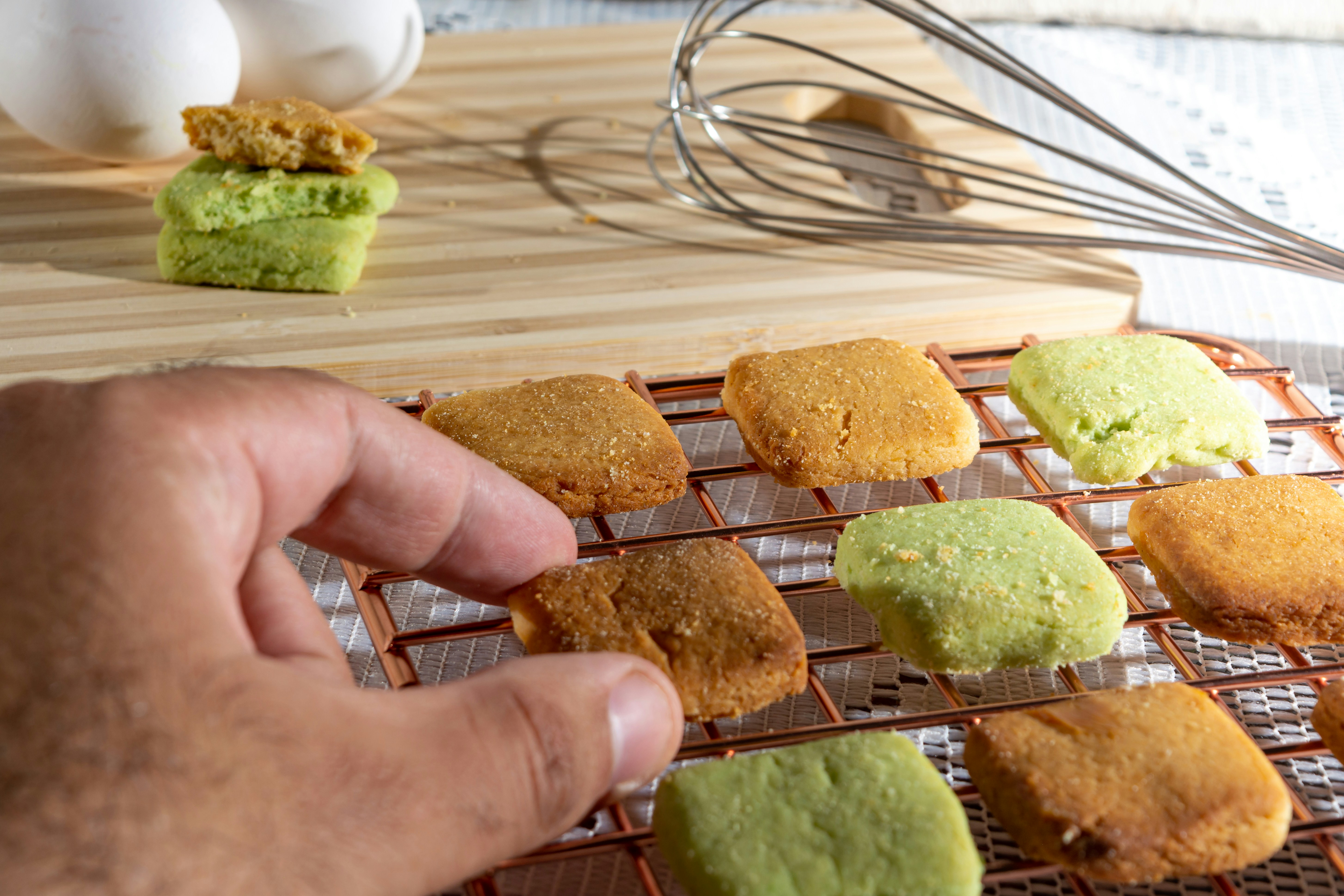 A hand reaches for a square cookie on a cooling rack. The cookies are golden brown and green in color. Behind the rack, a wooden board holds a stack of more cookies alongside a metal whisk, with some white eggs visible in the background.