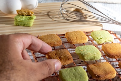 A hand reaches for a square cookie on a cooling rack. The cookies are golden brown and green in color. Behind the rack, a wooden board holds a stack of more cookies alongside a metal whisk, with some white eggs visible in the background.