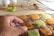 A hand reaches for a square cookie on a cooling rack. The cookies are golden brown and green in color. Behind the rack, a wooden board holds a stack of more cookies alongside a metal whisk, with some white eggs visible in the background.