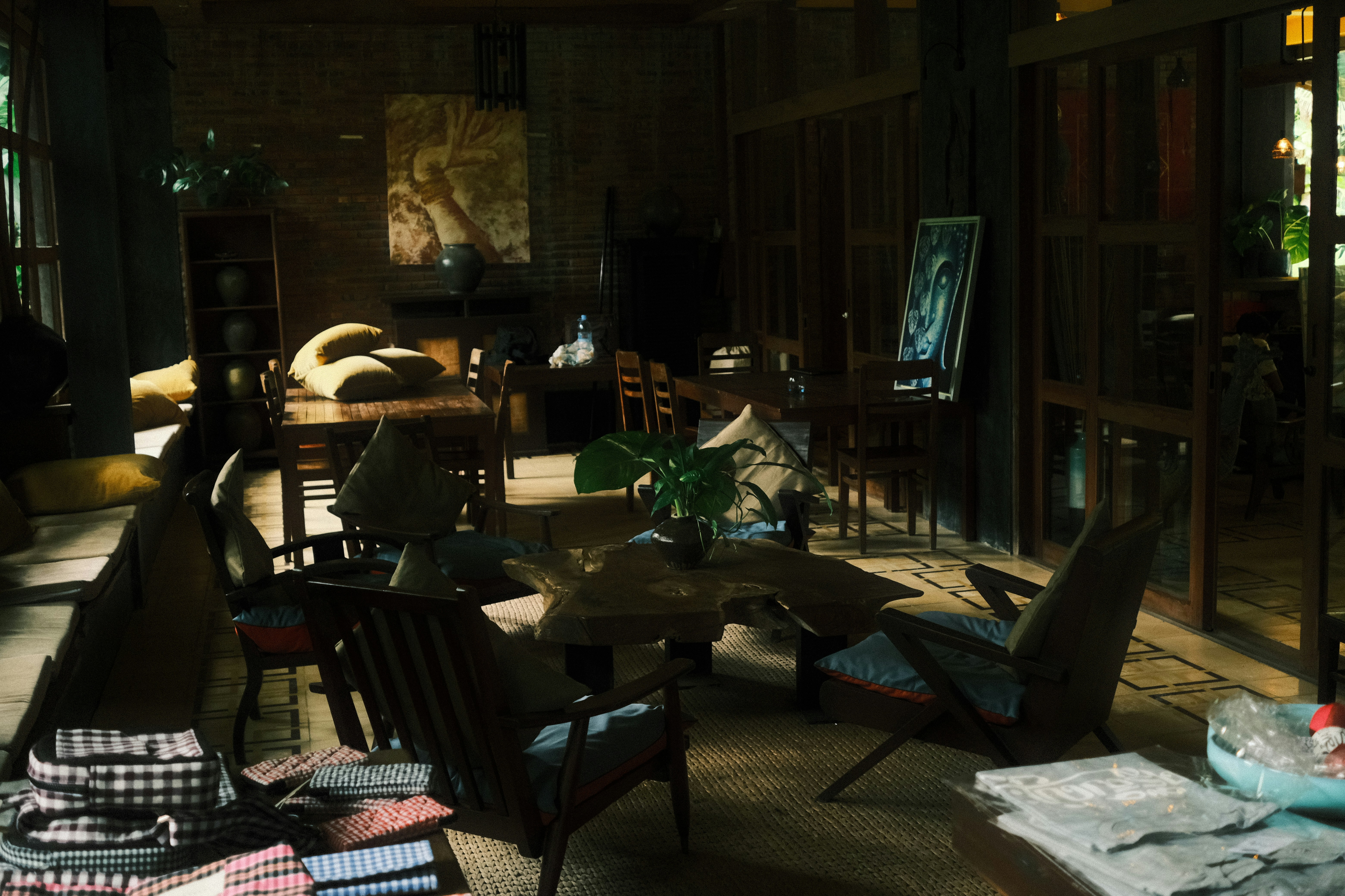 A cozy interior featuring wooden furniture and soft cushions. The room has a warm, inviting atmosphere with a large wooden table surrounded by chairs. Houseplants are placed throughout the room, adding a touch of nature. Soft lighting filters through large windows, casting gentle shadows across the space. A piece of art is displayed on an easel, contributing to the artistic ambiance.