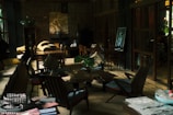 A cozy interior featuring wooden furniture and soft cushions. The room has a warm, inviting atmosphere with a large wooden table surrounded by chairs. Houseplants are placed throughout the room, adding a touch of nature. Soft lighting filters through large windows, casting gentle shadows across the space. A piece of art is displayed on an easel, contributing to the artistic ambiance.