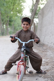 A young boy is joyfully riding a small, colorful tricycle on a dirt path. He is dressed in a traditional brown outfit and sandals, beaming with a wide smile. The background features a dirt wall and tall green trees, adding a natural setting to the scene.