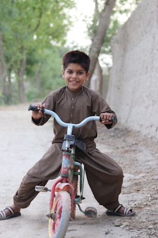 A young boy is joyfully riding a small, colorful tricycle on a dirt path. He is dressed in a traditional brown outfit and sandals, beaming with a wide smile. The background features a dirt wall and tall green trees, adding a natural setting to the scene.