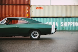 A classic dark green muscle car is parked on a wet pavement in front of stacked shipping containers. The vehicle features a sleek design with distinctive white racing stripes on the rear. The containers in the background are rust-colored and light teal, with industrial logos and the words 'CHINA SHIPPING' visible.
