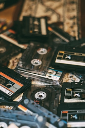 A collection of various audio cassette tapes scattered on a textured surface. The tapes have different designs, labels, and colors, primarily in shades of black, orange, and white. The image captures a vintage and nostalgic atmosphere with a focus on the physicality of analog music media.