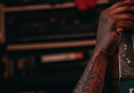 A close-up of a hand with tattoos resting on a textured surface, possibly holding or opening a box. The background is blurred, featuring shelves with electronic equipment or books, creating a dimly lit and cozy atmosphere.