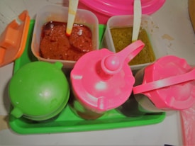 Two small containers with spoons are filled with red and green sauces or condiments, placed on a green tray along with two bottles with pink lids. The setup suggests a condiment serving area, possibly for adding flavor to food.