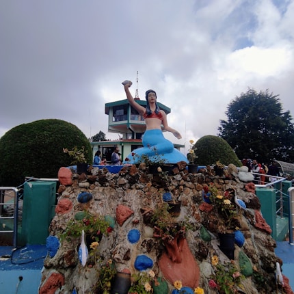 A statue of a mermaid appears prominently on a colorful, rock-covered pedestal adorned with small plants and flowers. The mermaid is holding a conch shell and is painted in vibrant colors, including a blue tail and a red top. Behind the statue is a building with large windows and a sign that reads 'TELESFERICO.' The sky is cloudy, with patches of blue peeking through.
