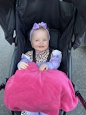 A smiling baby is sitting in a stroller, wearing a purple headband with a bow and a leopard print outfit. A bright pink blanket covers the lap. The background features a dark-colored stroller and a gray pavement.