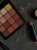 A makeup palette featuring various shades of brown and nude colors is laid out on a dark surface. Nearby, there is a tube of lipstick with a pinkish hue and a pair of small, silver geometric earrings shaped like triangles. A delicate silver chain with a small circular pendant is also visible.