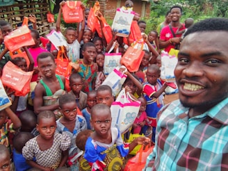 A large group of children and one adult are gathered in an outdoor setting. The children are smiling and holding colorful bags, predominantly orange and a few in white. The adult is taking a selfie and appears to be cheerful.