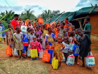 A diverse group of children and a few adults are gathered outdoors, holding brightly colored bags. They are standing in front of a small structure with a green tarp roof and mud walls. The children are wearing a variety of colorful clothes, and some are giving playful gestures.