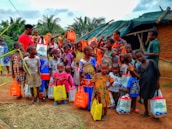 A diverse group of children and a few adults are gathered outdoors, holding brightly colored bags. They are standing in front of a small structure with a green tarp roof and mud walls. The children are wearing a variety of colorful clothes, and some are giving playful gestures.