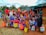 A diverse group of children and a few adults are gathered outdoors, holding brightly colored bags. They are standing in front of a small structure with a green tarp roof and mud walls. The children are wearing a variety of colorful clothes, and some are giving playful gestures.
