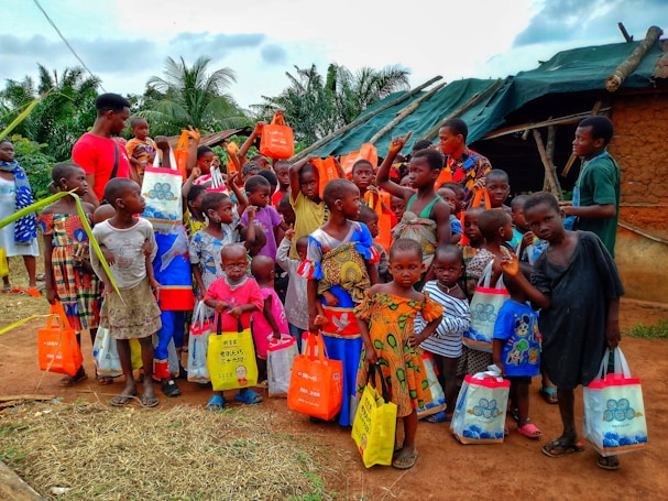 A diverse group of children and a few adults are gathered outdoors, holding brightly colored bags. They are standing in front of a small structure with a green tarp roof and mud walls. The children are wearing a variety of colorful clothes, and some are giving playful gestures.