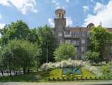 A multi-story residential building with a unique tower feature stands amidst lush green trees and a vibrant blue sky. In the foreground, there is an ornamental garden with manicured shrubs and a floral design on a grassy slope.