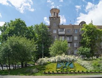 A multi-story residential building with a unique tower feature stands amidst lush green trees and a vibrant blue sky. In the foreground, there is an ornamental garden with manicured shrubs and a floral design on a grassy slope.