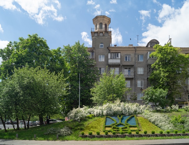 A multi-story residential building with a unique tower feature stands amidst lush green trees and a vibrant blue sky. In the foreground, there is an ornamental garden with manicured shrubs and a floral design on a grassy slope.