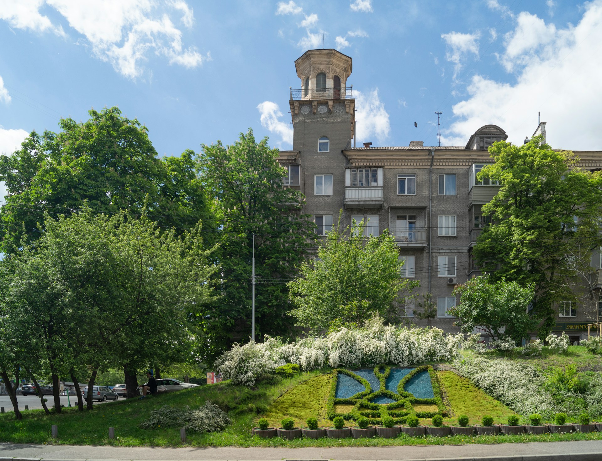 A multi-story residential building with a unique tower feature stands amidst lush green trees and a vibrant blue sky. In the foreground, there is an ornamental garden with manicured shrubs and a floral design on a grassy slope.