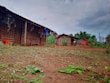 A rural setting with simple wooden structures lined up, featuring brick walls and corrugated metal roofs. Clothing hangs on lines strung across the buildings, with red containers positioned nearby. The ground is bare and earthy, scattered with green foliage. Tall green vegetation grows in the background under a cloudy sky.