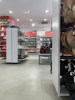 A shoe store with a variety of footwear on display, including sandals, formal shoes, and casual shoes. The shelves are neatly organized, and there is a section with handbags. The store has white shelving units and bright overhead lighting, with a red accent on the back wall displaying branded signage.