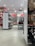 A shoe store with a variety of footwear on display, including sandals, formal shoes, and casual shoes. The shelves are neatly organized, and there is a section with handbags. The store has white shelving units and bright overhead lighting, with a red accent on the back wall displaying branded signage.
