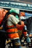 Workers clad in orange high-visibility vests and helmets are visible through a rain-soaked window, with raindrops creating a blurred effect. The background includes an out-of-focus street scene with signage and traffic.