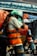 Workers clad in orange high-visibility vests and helmets are visible through a rain-soaked window, with raindrops creating a blurred effect. The background includes an out-of-focus street scene with signage and traffic.