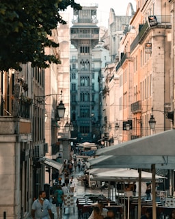 A bustling city street lined with historic buildings features an intricate architectural structure in the center. People are walking along the sidewalks, and several open-air cafes with umbrellas are visible. The street is vibrant with activity under a warm, soft lighting, while outdoor seating arrangements suggest a lively atmosphere.