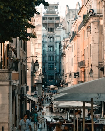 A bustling city street lined with historic buildings features an intricate architectural structure in the center. People are walking along the sidewalks, and several open-air cafes with umbrellas are visible. The street is vibrant with activity under a warm, soft lighting, while outdoor seating arrangements suggest a lively atmosphere.