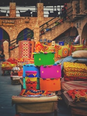 A vibrant display of colorful cushions and textiles is set against a rustic stone backdrop. The bright fabrics feature intricate patterns and bold colors, suggesting a lively market or artisan shop. The stone structure has arches and an outdoor staircase, adding to the traditional and historic ambiance.