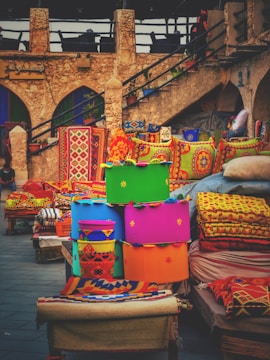 A vibrant display of colorful cushions and textiles is set against a rustic stone backdrop. The bright fabrics feature intricate patterns and bold colors, suggesting a lively market or artisan shop. The stone structure has arches and an outdoor staircase, adding to the traditional and historic ambiance.