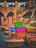 A vibrant display of colorful cushions and textiles is set against a rustic stone backdrop. The bright fabrics feature intricate patterns and bold colors, suggesting a lively market or artisan shop. The stone structure has arches and an outdoor staircase, adding to the traditional and historic ambiance.