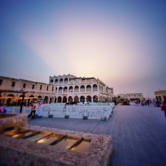 An open public square featuring a historic building with multiple arched windows and a white facade, surrounded by a white fence. People are walking and socializing in the vicinity, creating a lively atmosphere. The sky is clear with a warm gradient from blue to orange, suggesting a sunset or sunrise.