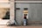 A person stands at an ATM on a city street, interacting with it. A bicycle is parked nearby against a brick building that features a large window showing indoor plants and furniture.